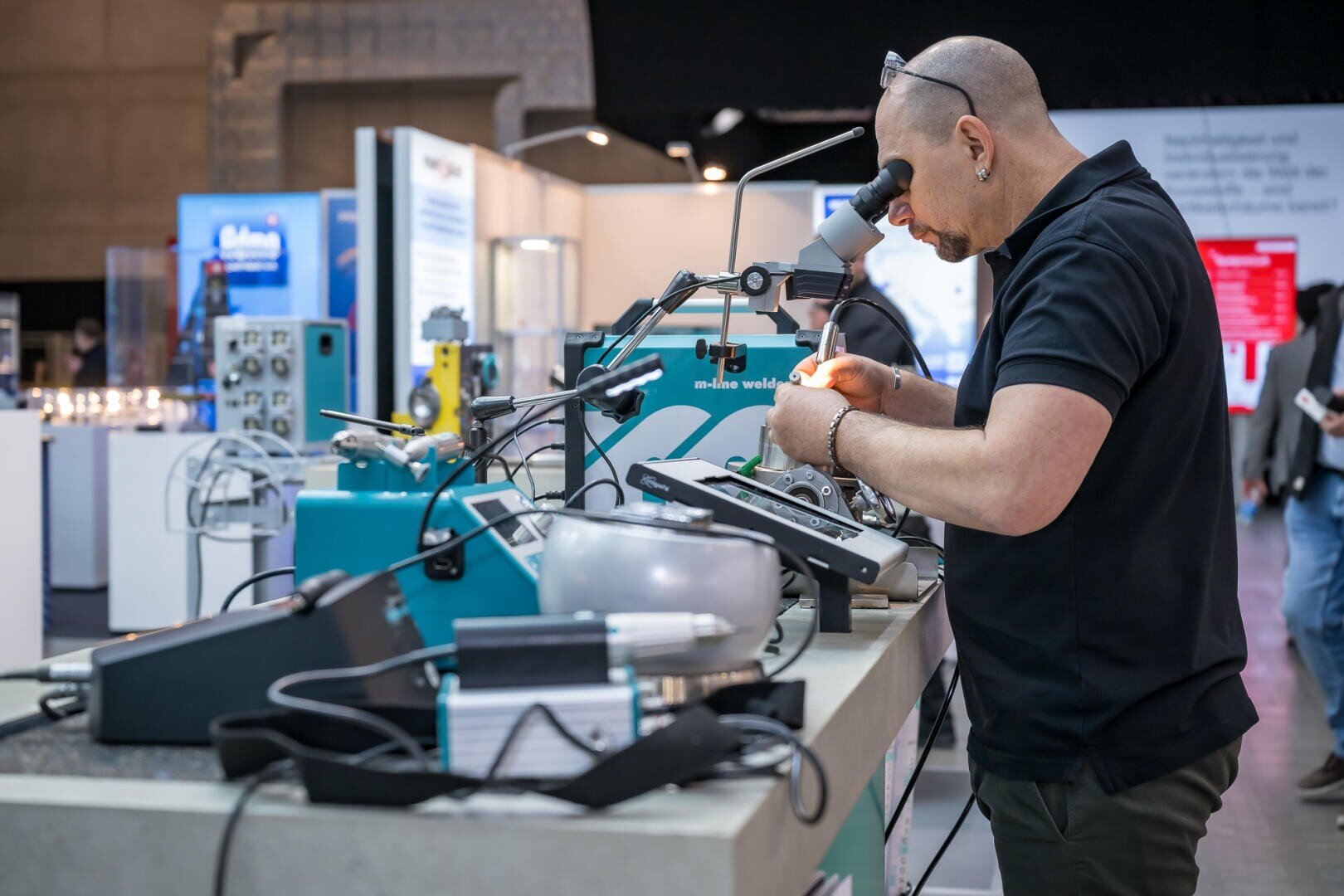 Eine Person mit Brille arbeitet an einer Werkbank mit einem Mikroskop und Lötwerkzeugen und konzentriert sich auf komplizierte elektronische Bauteile. Auf der Theke eines modernen Labors sind verschiedene technische Geräte und Kabel ausgebreitet.