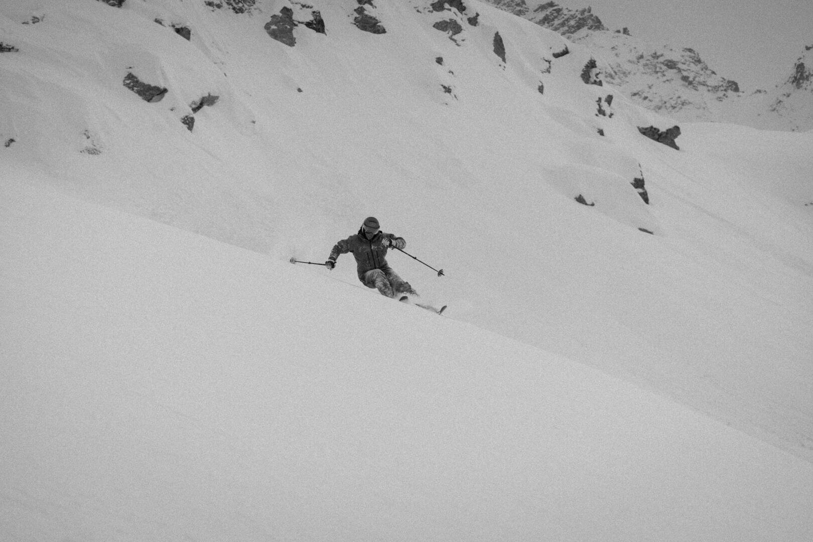 Ein Skifahrer fährt einen verschneiten, steilen Berghang hinunter, umgeben von Felsen und zerklüftetem Gelände, aufgenommen in Schwarz-Weiß.