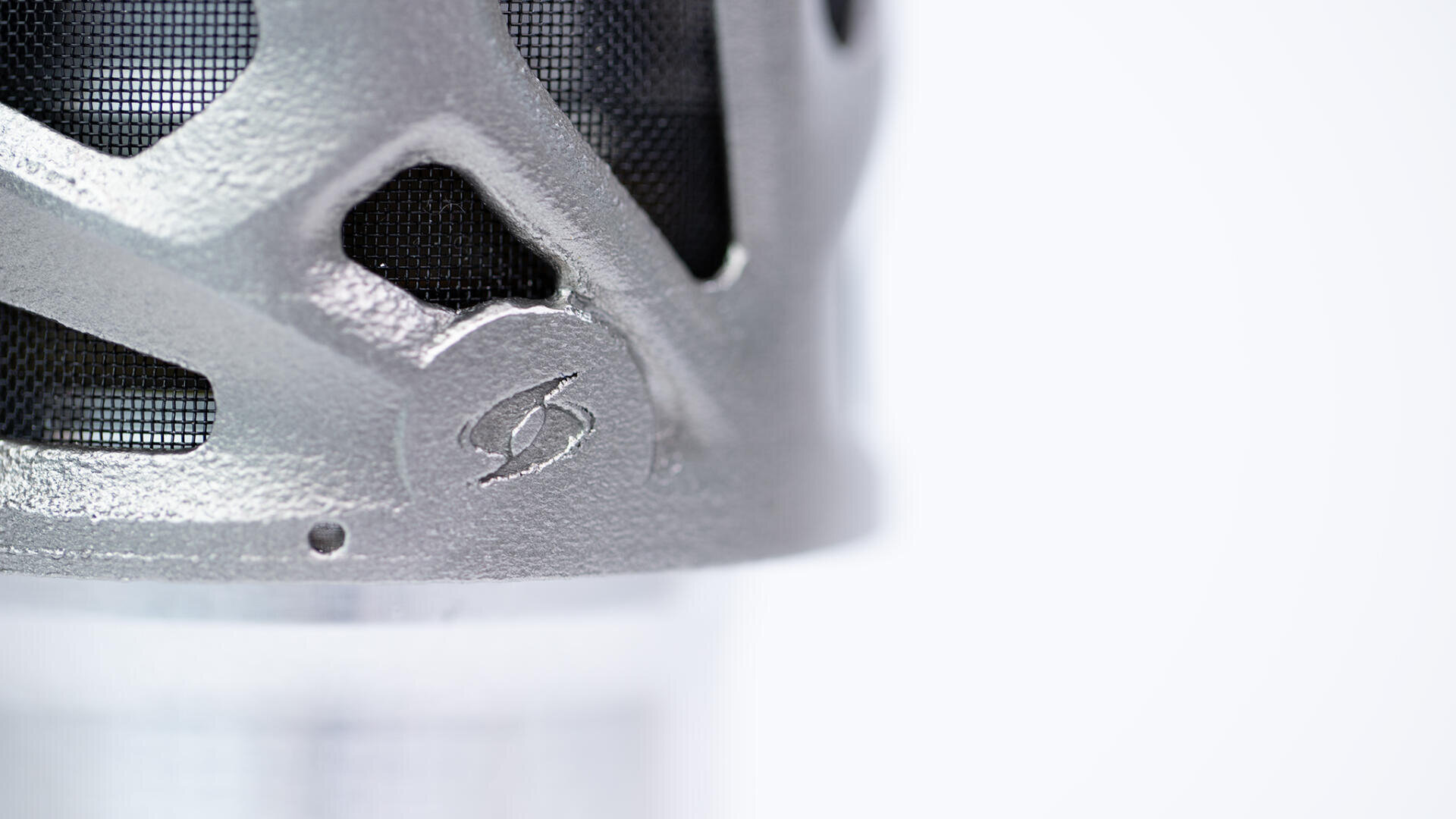 Nahaufnahme eines metallischen Objekts mit einem Maschendrahtgitter und einer geometrischen Struktur, auf dessen Oberfläche ein Blitzsymbol eingraviert ist, vor einem schlichten weißen Hintergrund.