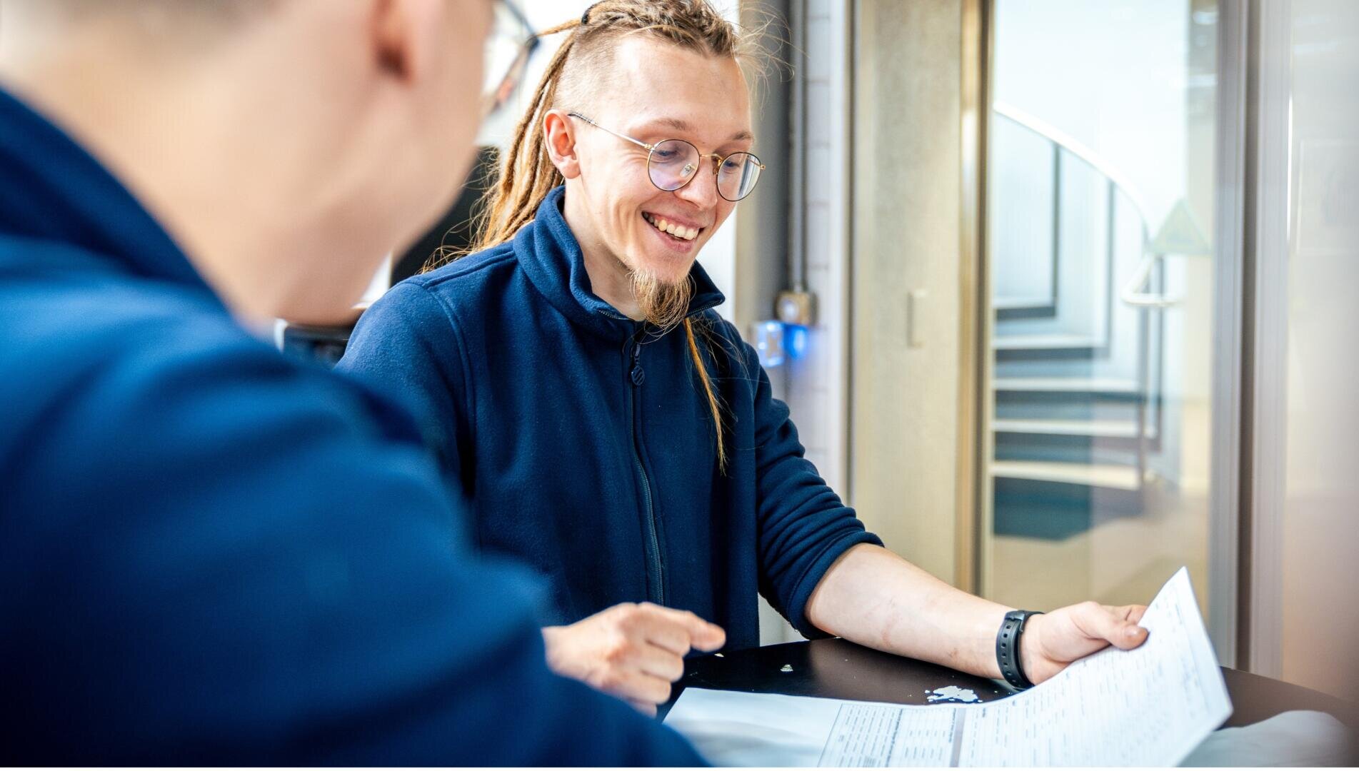 Eine lächelnde Person mit Brille und langen Haaren in einer blauen Jacke sitzt an einem Tisch, hält Papiere in der Hand und unterhält sich mit einer anderen Person in einer Büroumgebung.