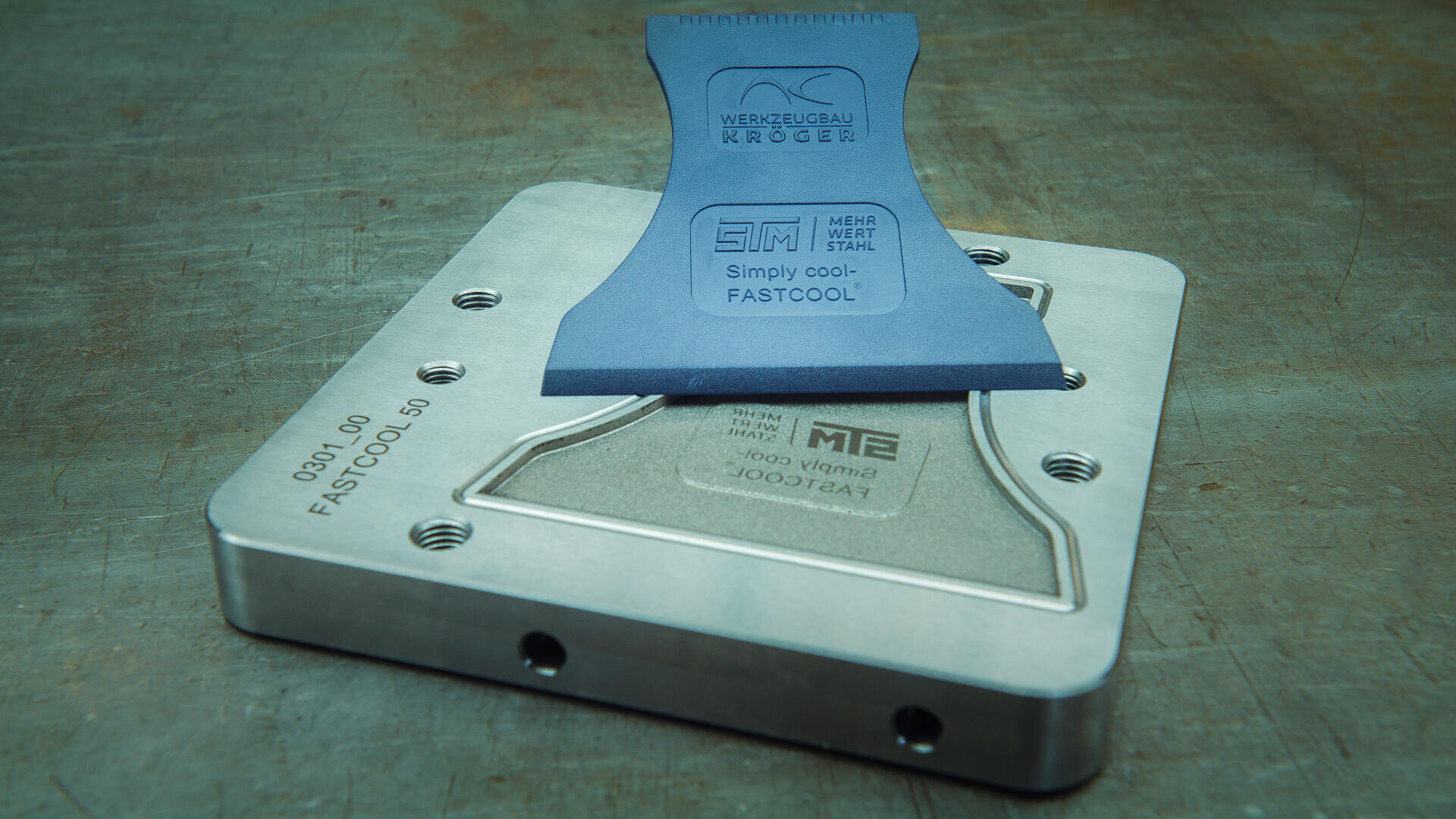 Un outil en plastique bleu portant l'inscription Simply cool-FASTCOOL repose sur une pièce métallique rectangulaire avec un texte gravé, des vis et une surface brossée, le tout sur un fond métallique.