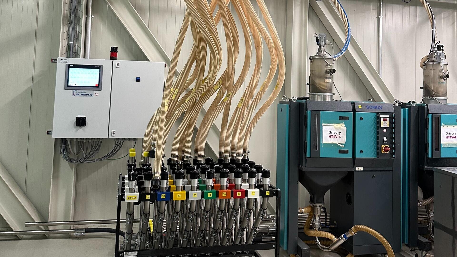 Eine Reihe von Industriemaschinen mit flexiblen Schläuchen, die mit einer Schalttafel und zwei großen, blaugrünen Einheiten verbunden sind und für die Verarbeitung oder den Transport von Materialien in einer Produktionsanlage verwendet werden.