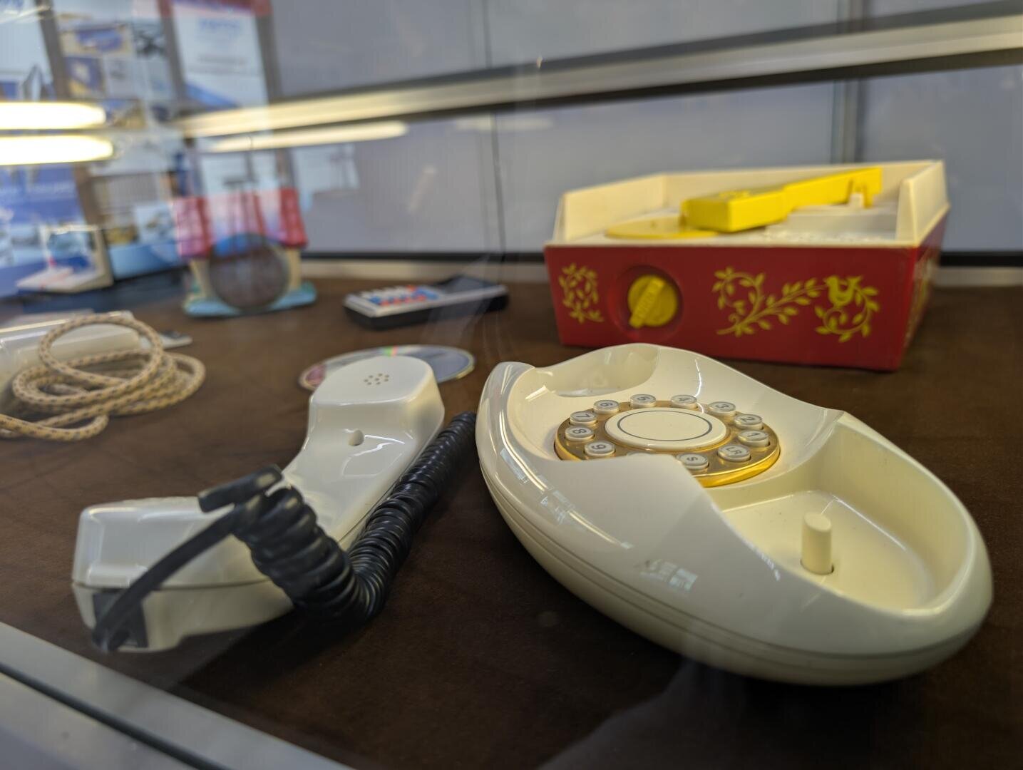 Eine Vitrine mit einem alten weißen Wählscheibentelefon, einer roten und gelben Schachtel, einem aufgerollten Seil und anderen Retro-Artikeln, die auf einer braunen Fläche angeordnet sind.