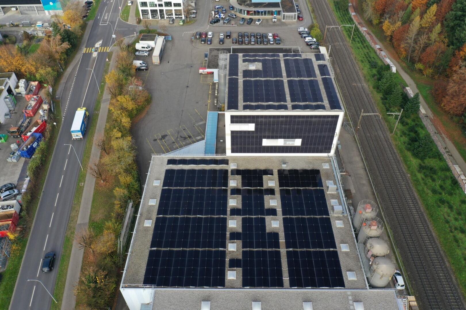 Luftaufnahme eines Gebäudes mit mehreren großen Solarmodulen auf dem Dach, neben einer Bahnlinie, einem Parkplatz und umliegenden Straßen mit Fahrzeugen und Bäumen in Herbstfarben.