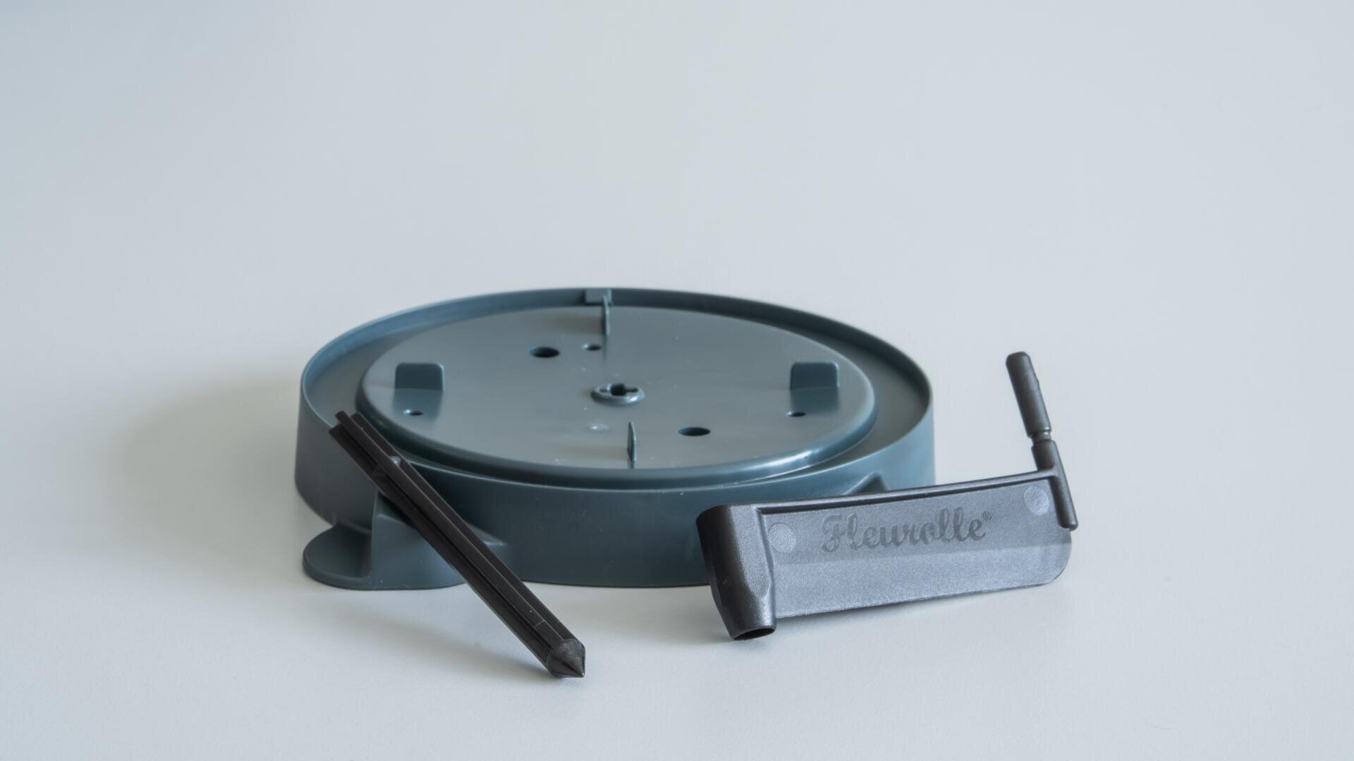 Ein runder, grauer Kunststoffsockel mit erhabenen Stiften, ein schwarzes Kunststoffwerkzeug und ein grauer Clip mit der Aufschrift Flowelle auf einer schlichten weißen Oberfläche.