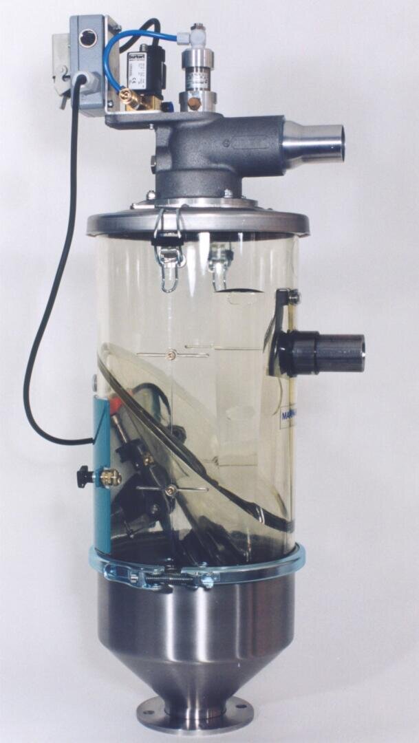 Ein transparentes zylindrisches Industriegerät mit Metallbeschlägen, Drähten und mechanischen Teilen, einschließlich einer oberen Düse, eines Schaltkastens und eines konischen Metallsockels, alles vor einem schlichten hellen Hintergrund.