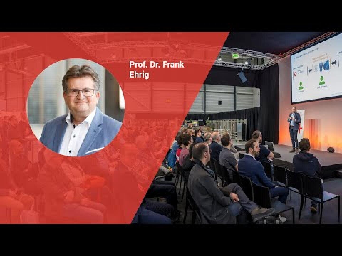 Un homme en costume apparaît en surimpression rouge avec le texte Prof. À l'arrière-plan, il s'adresse à un public dans une grande salle de conférence équipée d'un écran de projection.