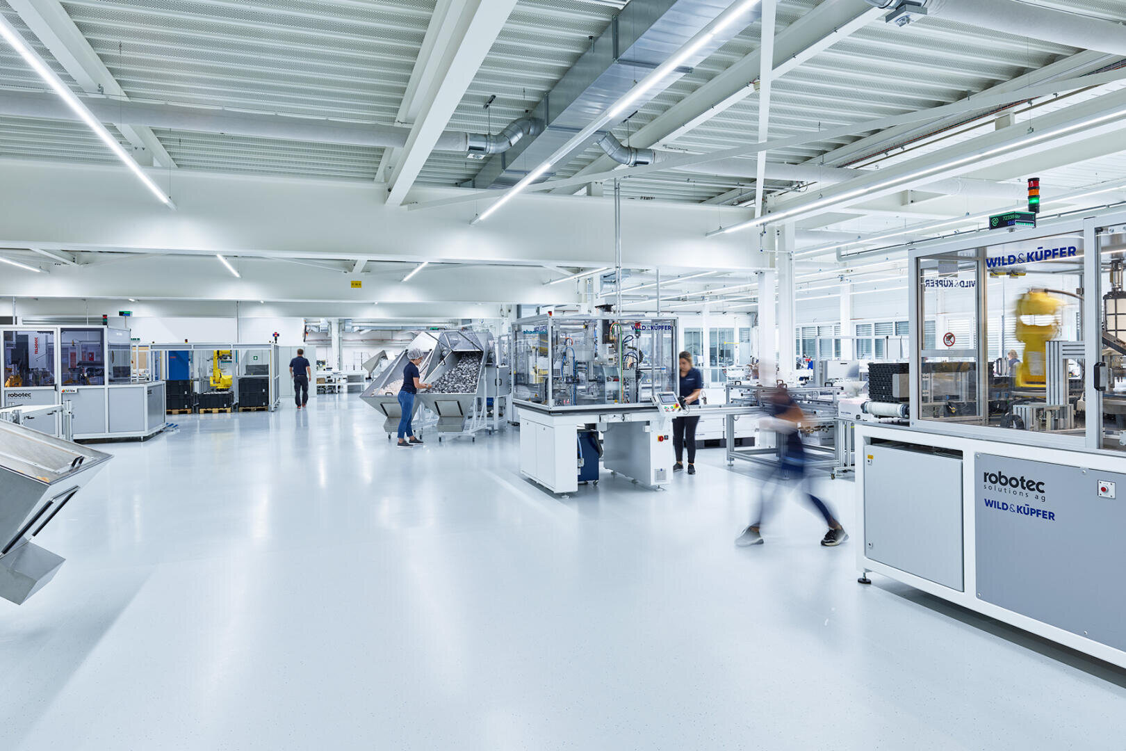 Un hall d'usine moderne et lumineux où les gens travaillent entre de grandes machines et installations industrielles, y compris des systèmes robotisés. L'espace est propre, spacieux et bien éclairé avec des sols et des plafonds blancs.