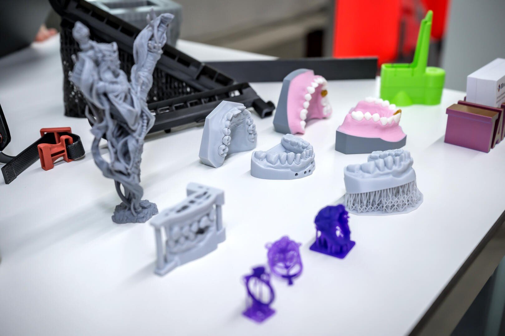 Une multitude de modèles de dents, de prototypes et de structures imprimés en 3D dans les tons gris, rose, vert et violet sont exposés sur une table blanche dans un environnement de laboratoire.