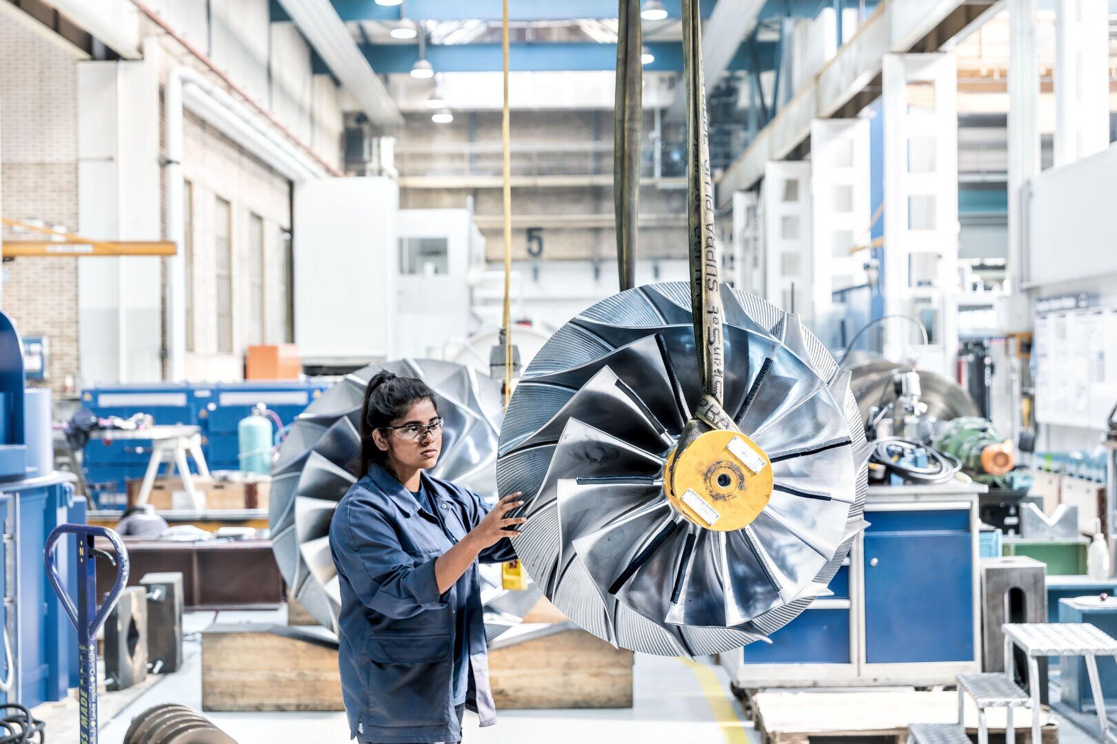 Eine Person mit Schutzbrille und blauem Laborkittel inspiziert eine große industrielle Turbinenschaufel, die in einer mit Maschinen und Geräten gefüllten Werkstatt aufgehängt ist.