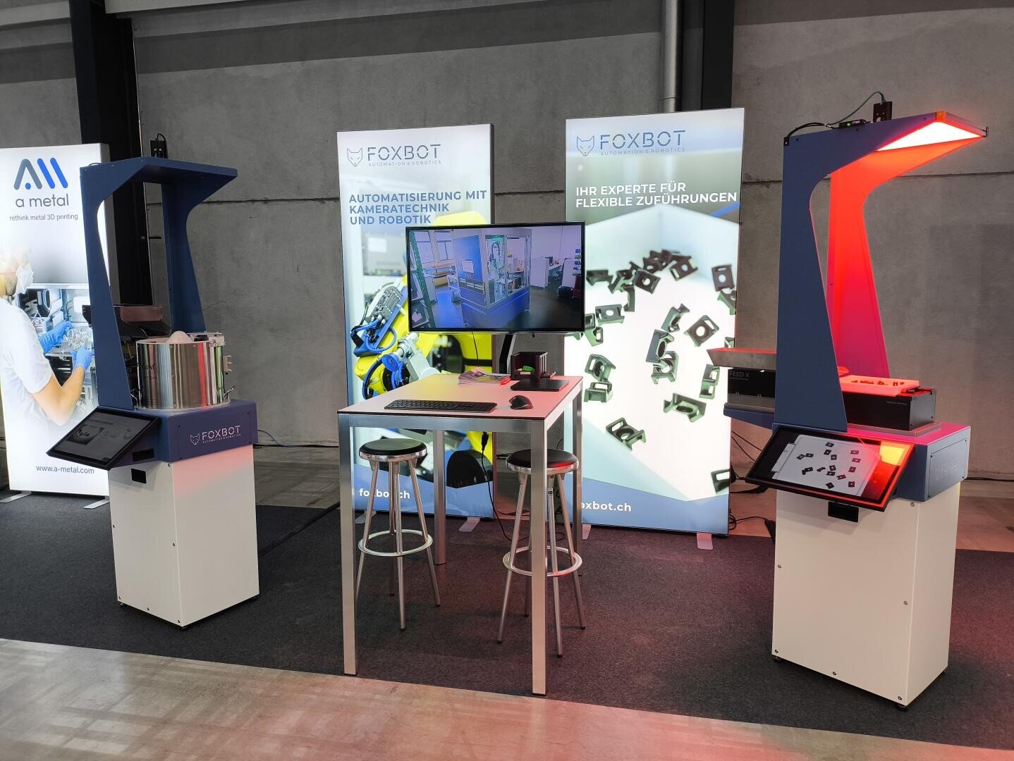 Ein Messestand präsentiert die FOXBOT-Roboterautomation mit zwei Scanstationen, einem zentralen Tisch mit Hockern, einem Monitor, der Bilder anzeigt, und Bannern in deutscher Sprache, die für flexible Zuführlösungen werben.