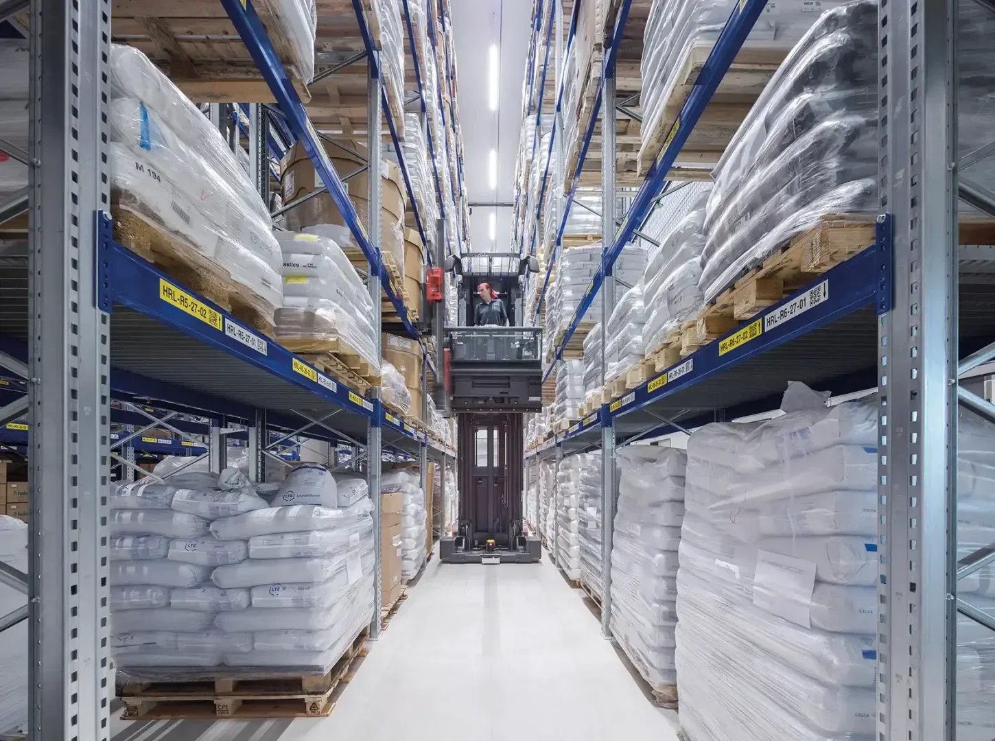 Un ouvrier manie un chariot élévateur dans une allée d'entrepôt entourée de hauts rayonnages où s'empilent de grands paquets blancs et des caisses sur des palettes en bois. L'environnement est bien éclairé et ordonné.