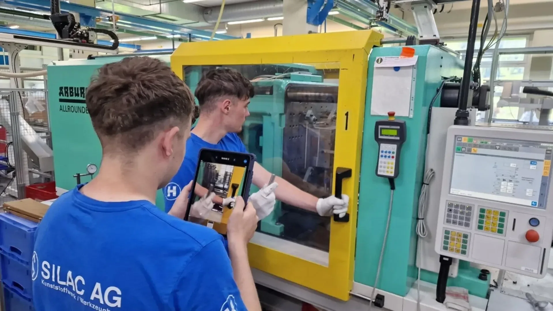 Zwei junge Männer bedienen und überprüfen eine große Industriemaschine in einer Fabrik. Der eine fotografiert oder zeichnet mit einem Tablet auf, während der andere mit Handschuhen an der Maschine hantiert. Beide tragen blaue Hemden der SILAC AG.