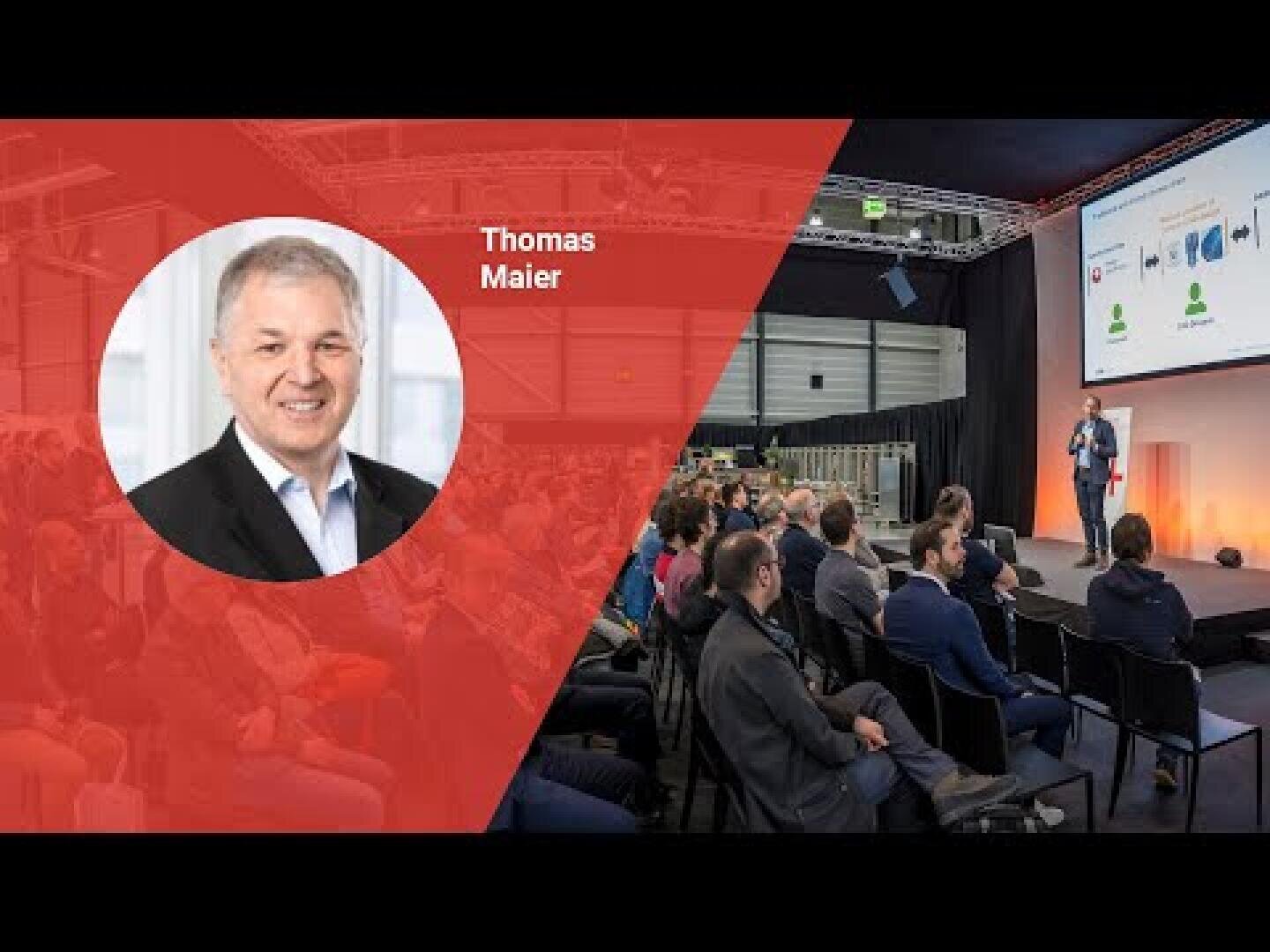 Un homme en costume se tient sur scène et donne une conférence devant un public assis. Sur le côté gauche, une superposition rouge montre la photo d'un homme souriant avec l'inscription Thomas Maier. Le lieu de la manifestation semble moderne et bien éclairé.