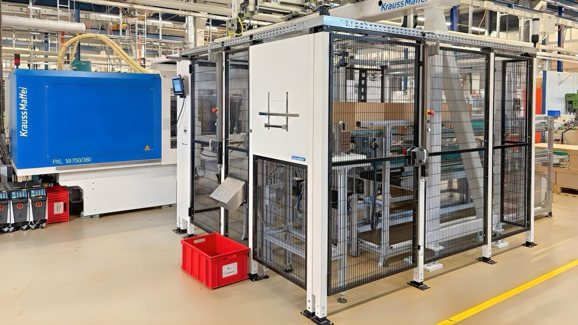 Une machinerie industrielle moderne dans une usine avec un grand appareil KraussMaffei bleu, un boîtier en métal et en verre et un réservoir en plastique rouge sur le sol, entouré de divers tuyaux et appareils.