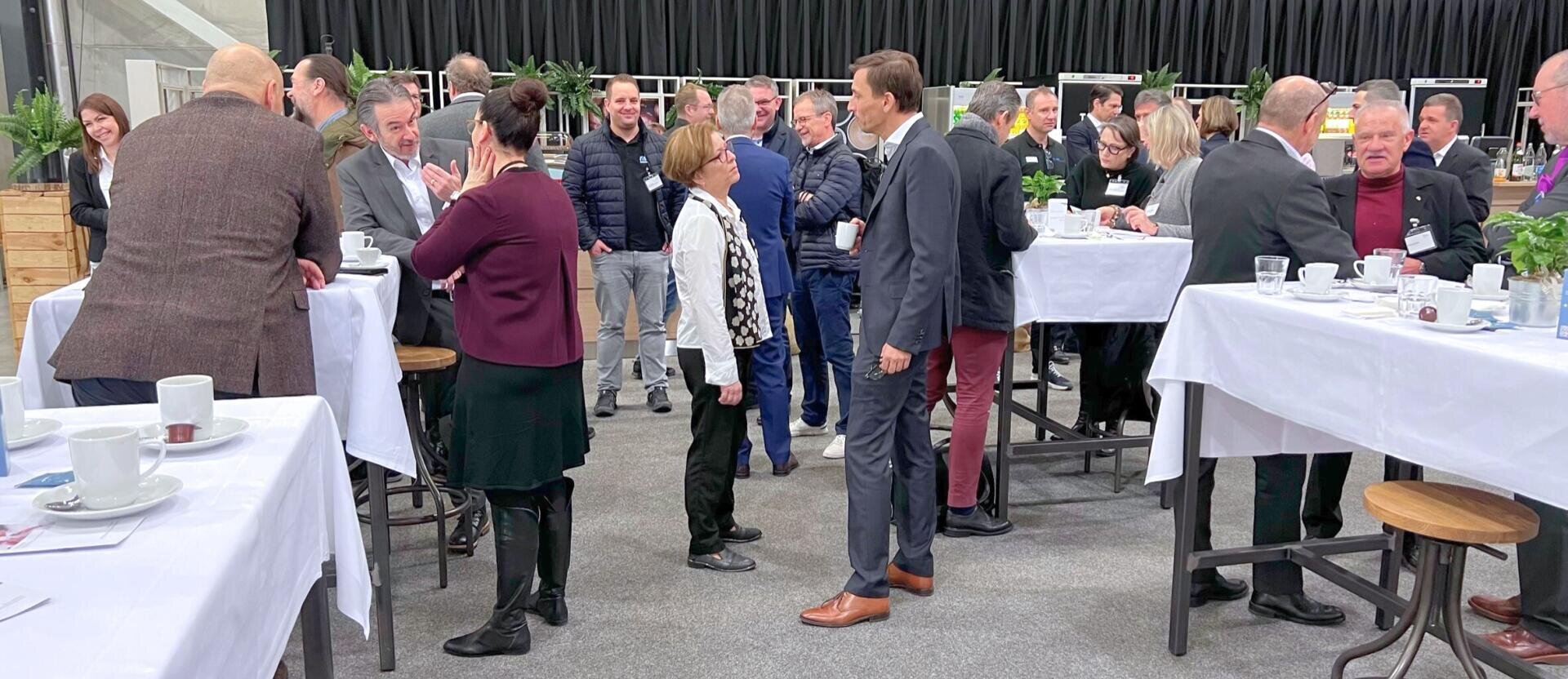 Eine Gruppe von Menschen knüpft bei einer Indoor-Veranstaltung an Stehtischen mit Kaffeetassen Kontakte. Einige stehen und unterhalten sich, während andere sitzen oder sich zwischen den Tischen bewegen. Die Atmosphäre ist lebhaft und locker.