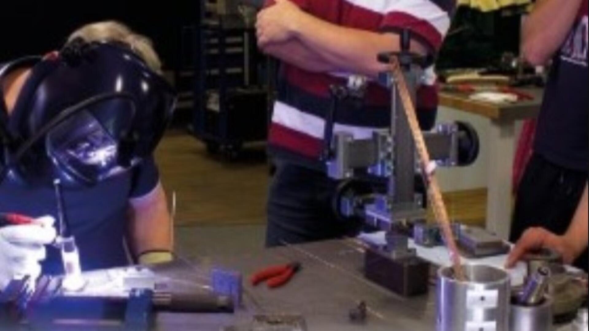 Eine Person mit einem Schweißhelm arbeitet an einem Tisch mit Werkzeugen und Metallgeräten, während zwei weitere Personen in der Nähe stehen und beobachten. Verschiedene Werkzeuge und mechanische Teile sind auf dem Tisch verstreut.