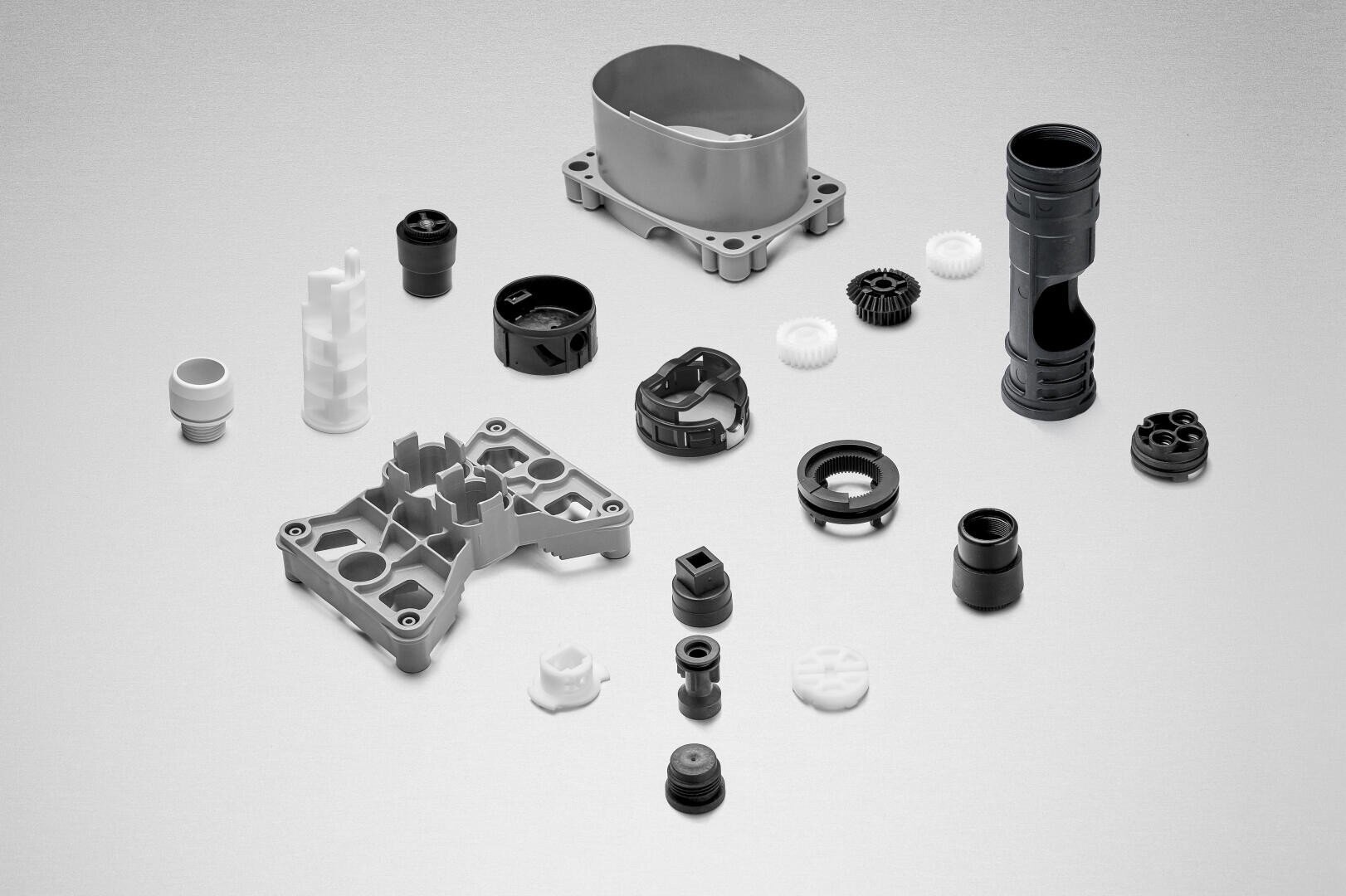 Une collection de diverses pièces mécaniques en plastique et en métal, y compris des engrenages, des boîtiers et des connecteurs, disposées proprement sur une surface gris clair.