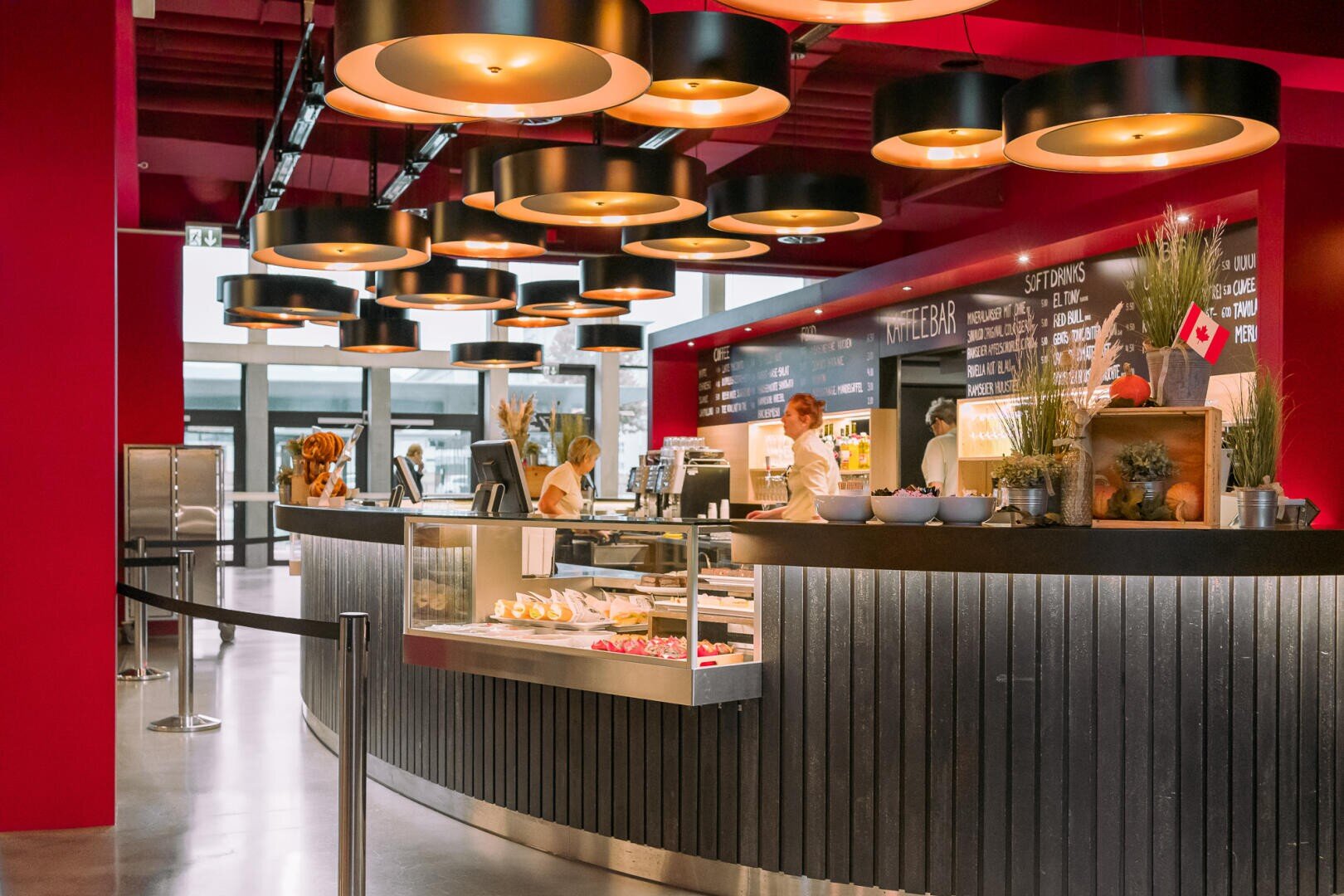 Une cafétéria moderne avec des murs rouges et des lampes rondes suspendues. Le personnel derrière le comptoir prépare les repas, tandis que des pâtisseries et des boissons sont proposées. Des tableaux de menus sont visibles en arrière-plan. L'espace est propre et accueillant.