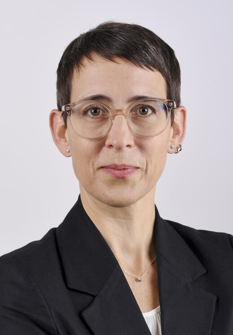 Une personne aux cheveux foncés et courts, portant des lunettes claires, un blazer noir, un collier clair et de petites boucles d'oreilles, regarde avec assurance l'appareil photo sur un fond clair et sobre.