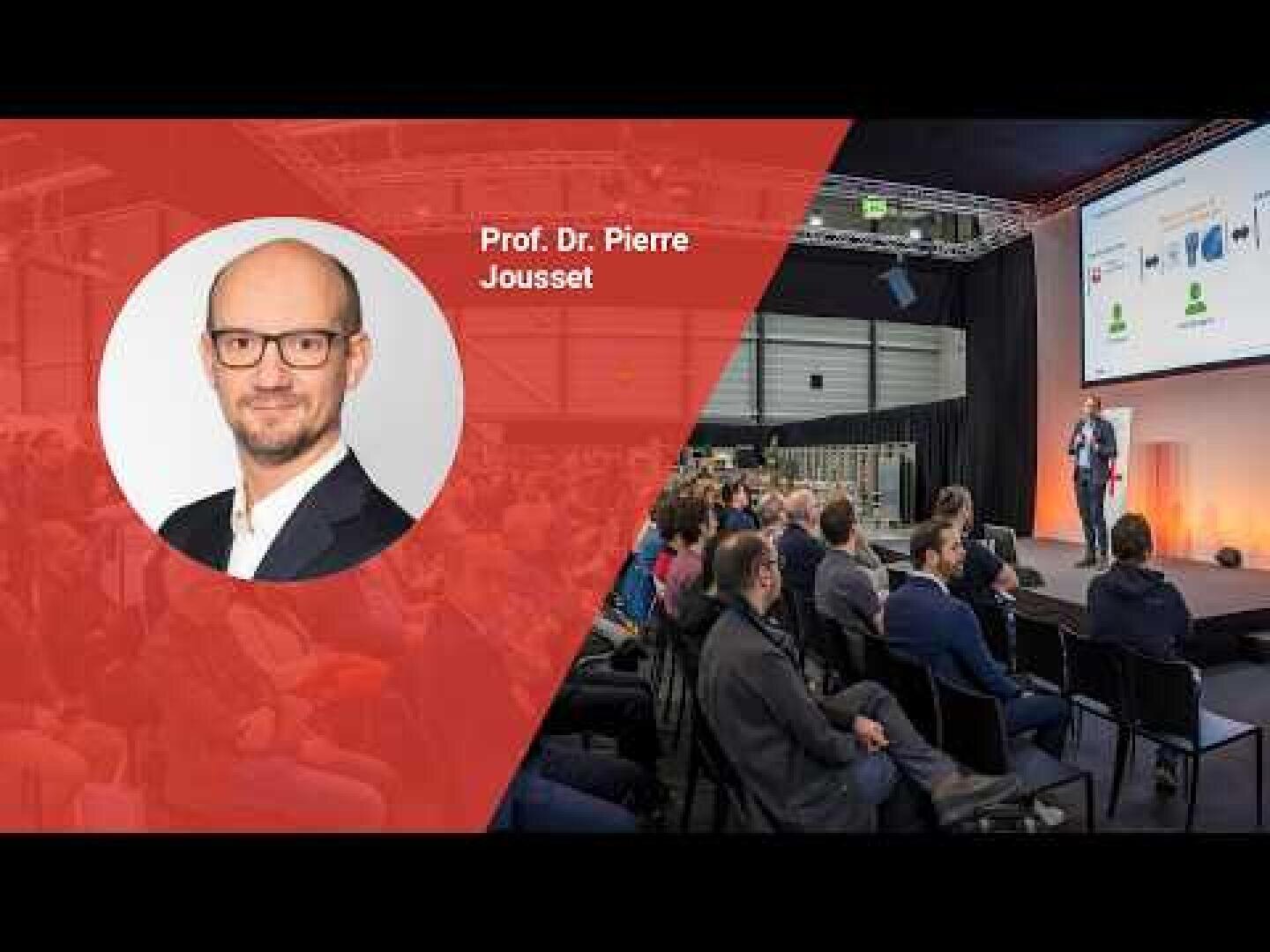 Eine Präsentationsszene mit dem Foto und dem Namen von Prof. Dr. Pierre Joussets auf einem roten, transparenten Overlay, während das Publikum einem Redner zuhört, der auf der Bühne eines modernen Konferenzsaals Folien präsentiert.