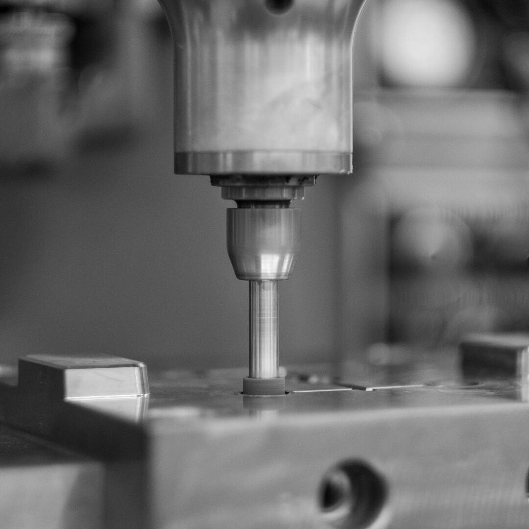 Un gros plan en noir et blanc d'une fraiseuse CNC qui découpe ou perce une pièce de métal, avec un focus sur l'outil en rotation et des machines floues en arrière-plan.