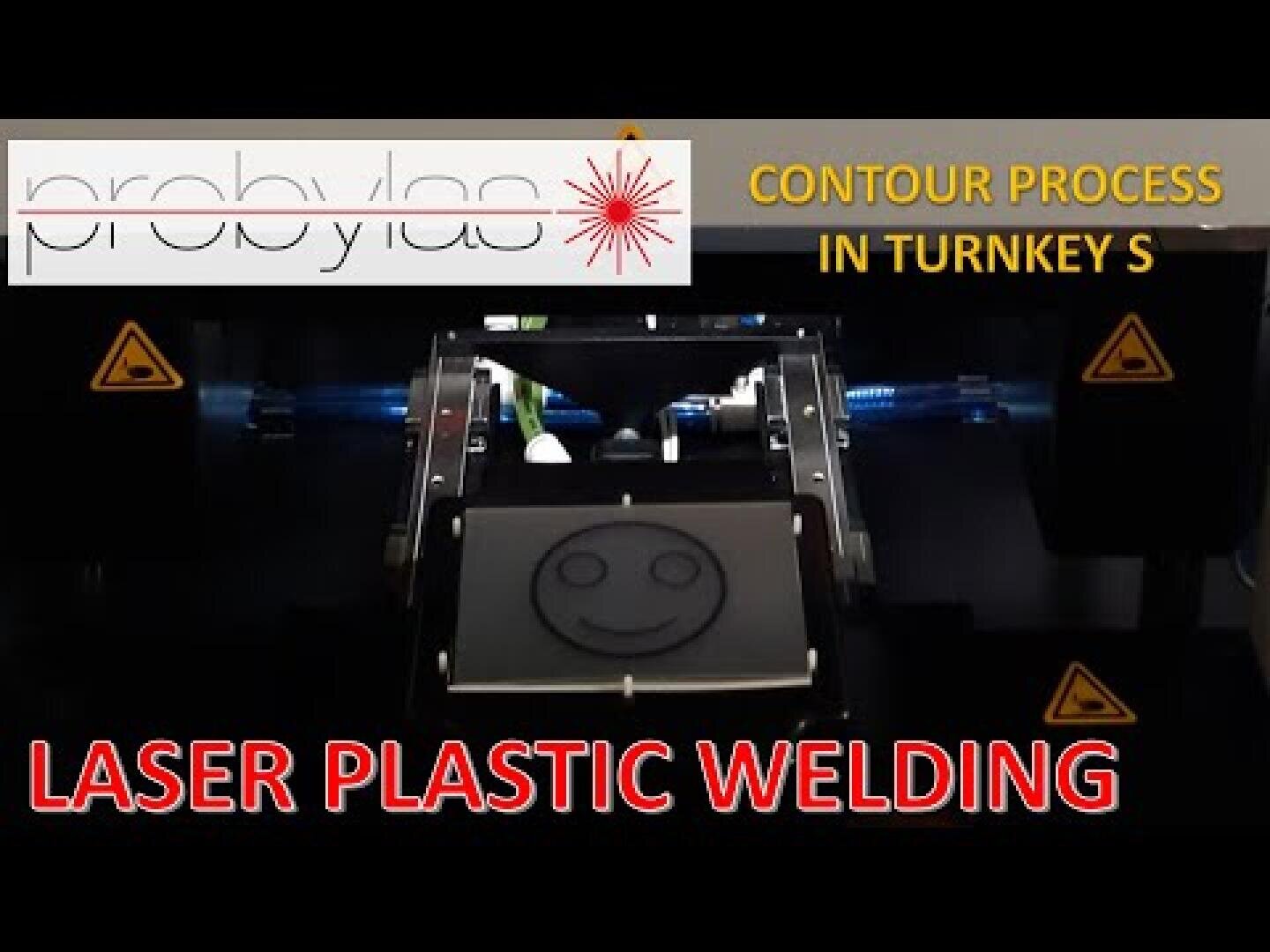 Eine Maschine, die das Laserschweißen von Kunststoffen durchführt, mit einem Smiley-Gesicht auf dem Kunststoff. Der Text lautet probylas CONTOUR PROCESS IN TURNKEY S und LASER PLASTIC WELDING. Auf beiden Seiten sind Warnschilder zu sehen.