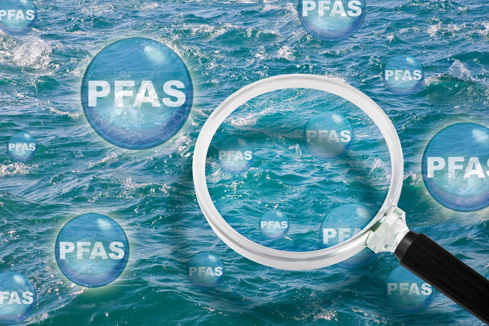 Ein Vergrößerungsglas schwebt über blauem Meerwasser und hebt transparente Blasen mit der Aufschrift PFAS hervor, die den Nachweis von PFAS-Chemikalien im Wasser symbolisieren.