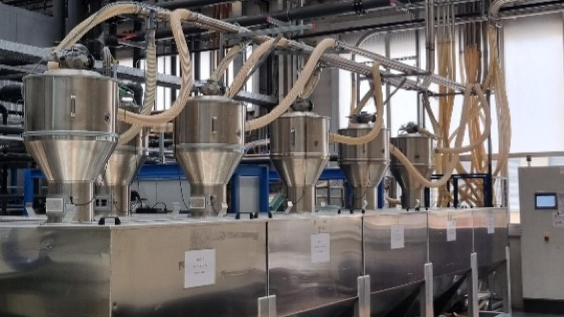 Reihen von Industrietrichtern aus Edelstahl, die durch transparente Rohre miteinander verbunden sind, in einer modernen Fabrik, mit Rohren und Schalttafeln im Hintergrund.