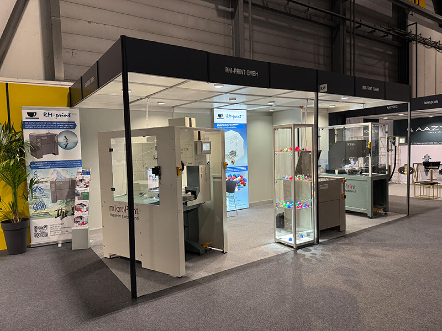 Stand d'exposition de RM-Print GmbH avec de grandes machines industrielles, des vitrines avec des objets imprimés en 3D aux couleurs vives, des bannières avec des informations sur l'entreprise et une structure propre et moderne dans une salle de congrès.