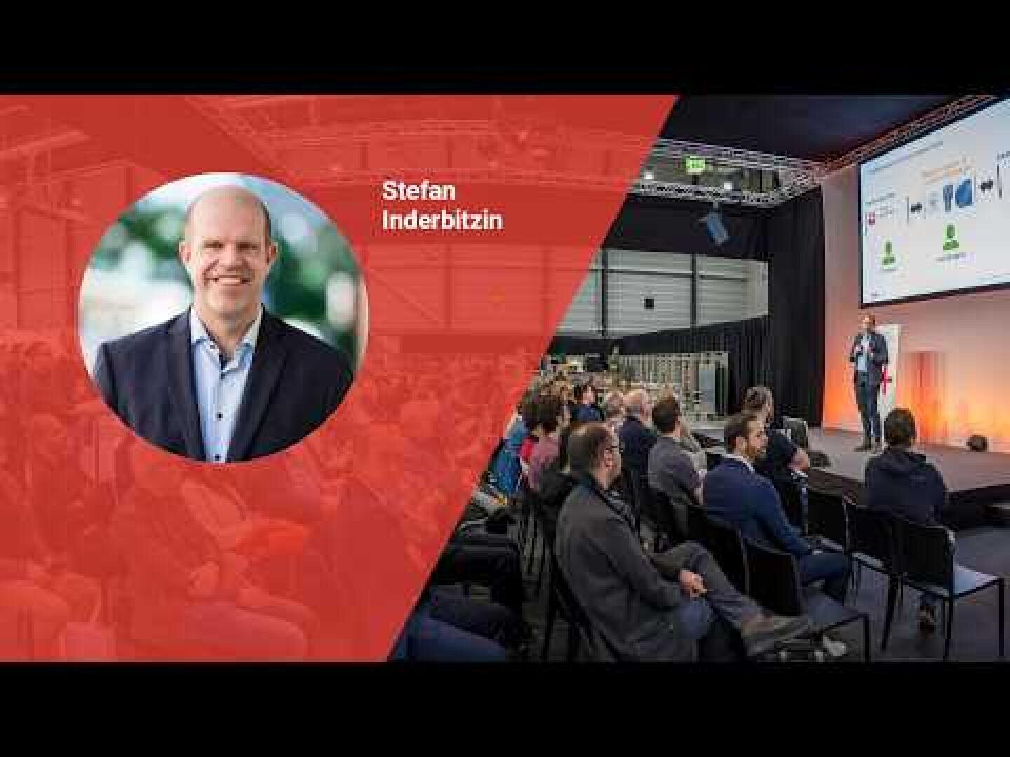 Ein Mann, der als Stefan Inderbitzin identifiziert wird, spricht in einem Konferenzraum zu einem Publikum. Auf der linken Seite ist sein Porträt in einer roten Grafik eingerahmt, daneben wird sein Name angezeigt.