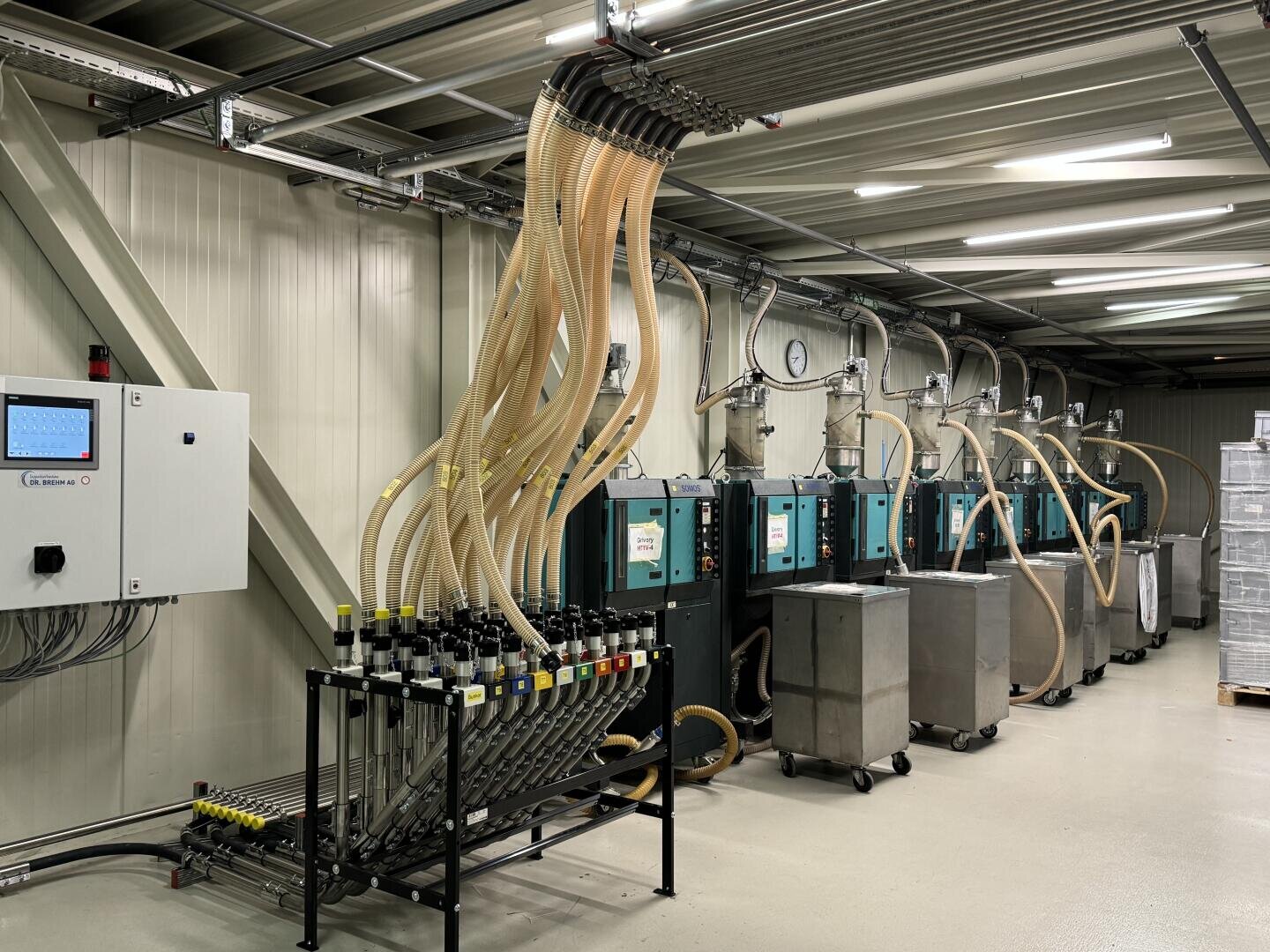 Industrieraum mit mehreren großen Maschinen, die durch transparente Schläuche mit einer Schalttafel und einem Verteiler verbunden sind; die Schläuche sind von der Decke abgehängt und an jeder Maschine für den Flüssigkeits- oder Materialtransport angebracht.