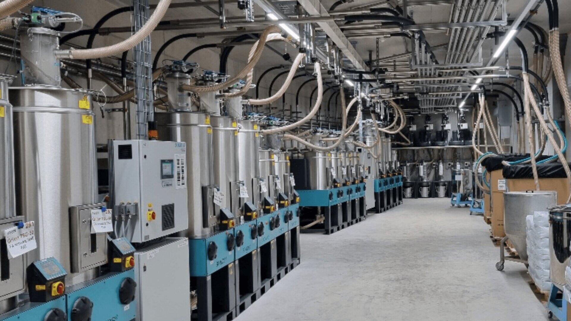 Une salle d'usine remplie de machines industrielles, de tuyaux, de panneaux de contrôle et de câbles qui courent au-dessus de la tête. L'équipement est organisé en rangées de part et d'autre d'une allée large et propre.