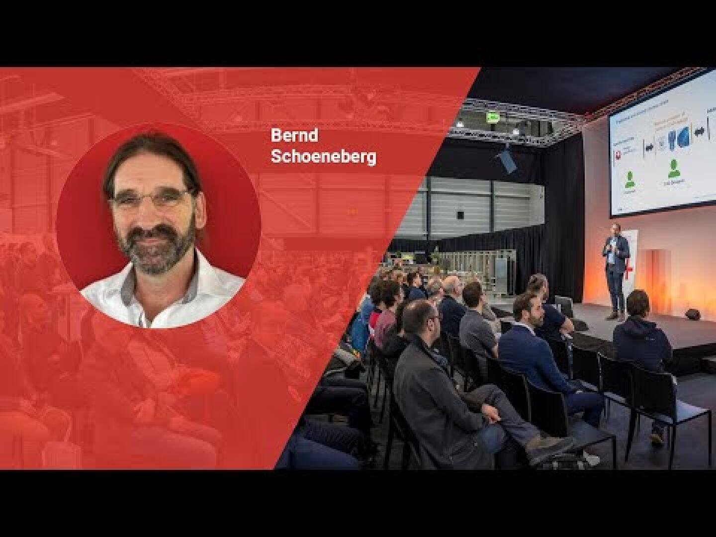 Ein Redner spricht auf der Bühne zu einem Publikum. Auf der linken Seite zeigt eine überlagerte rote Grafik das Foto eines Mannes mit Brille und Bart, der Bernd Schoeneberg heißt.