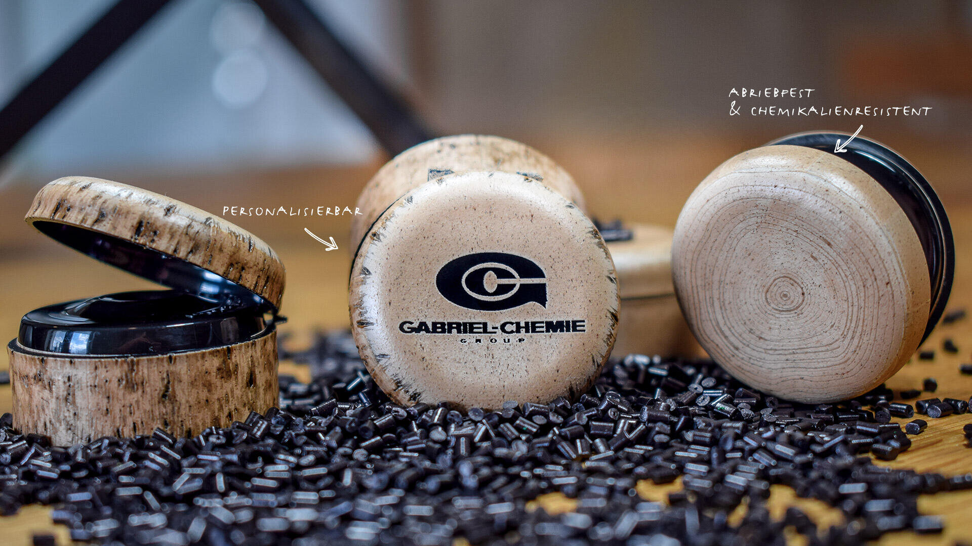 Drei runde Kosmetikbehälter mit Deckeln, von denen einer geöffnet ist und ein schwarzes Inneres zeigt, befinden sich zwischen verstreuten schwarzen Kügelchen. Der mittlere Behälter trägt ein Gabriel-Chemie-Logo. Handgeschriebene Etiketten weisen auf PERSONALISIERBAR und ADELKAPPE & CHEMIKALIENBESTÄNDIG hin.