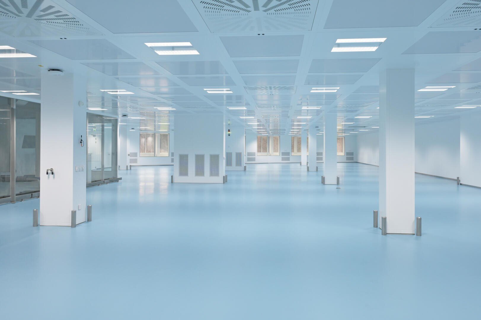 Ein großer, leerer, moderner Raum mit hellblauem Boden, weißen Wänden und Deckenleuchten. Säulen sind im ganzen Raum verteilt, und Glastüren und Fenster säumen Teile der Wände, was dem Raum einen sauberen, sterilen Eindruck verleiht.