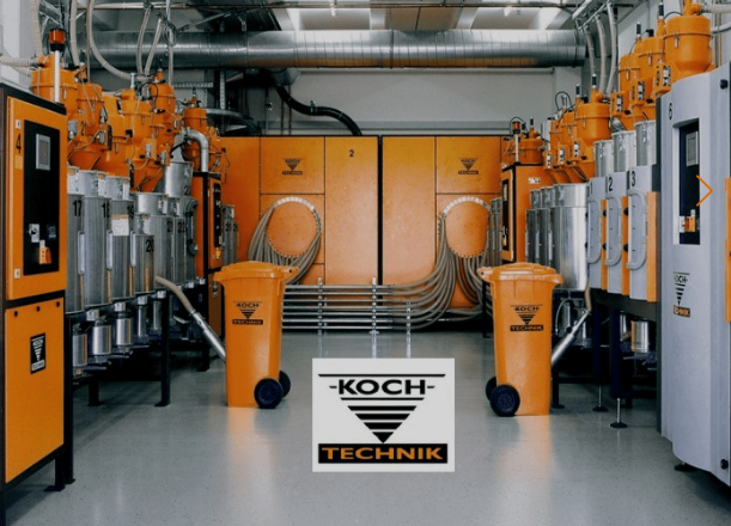 Industrieanlage mit orangefarbenen und silbernen Maschinen, Behältern und Geräten, die symmetrisch angeordnet sind. Das große KOCH TECHNIK-Logo ist mittig im Vordergrund zu sehen. Rohre und Kanäle verlaufen an der Decke und den Wänden.