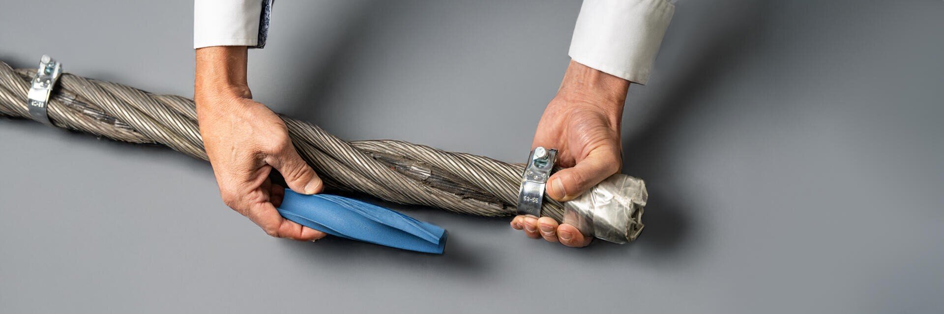 Die Hände einer Person halten ein dickes, verdrilltes Metallkabel und ziehen eine blaue Schutzhülle darüber; das Kabel ist an den Enden mit Klemmen und Folie versehen. Der Hintergrund ist eine einfarbig graue Fläche.