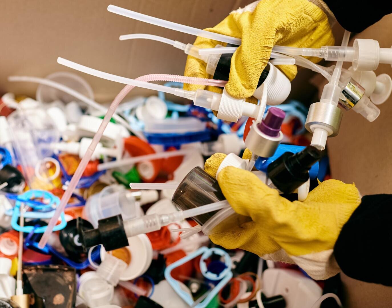 Eine Person mit gelben Gummihandschuhen sortiert verschiedene Pump- und Sprühflaschenverschlüsse aus Kunststoff über einer Kiste, die mit gemischten Kunststoffabfällen und Wertstoffen gefüllt ist.