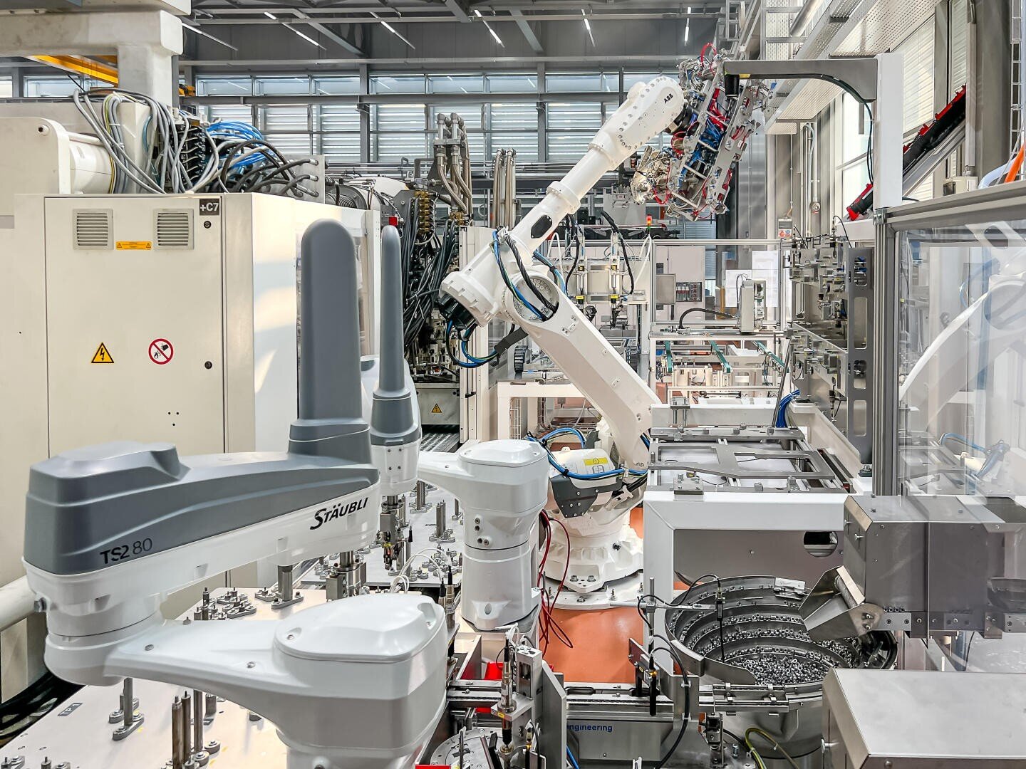 Une usine moderne avec plusieurs bras robotisés et des machines automatisées qui assemblent des produits dans un environnement industriel lumineux avec de grandes fenêtres et différentes pièces métalliques.