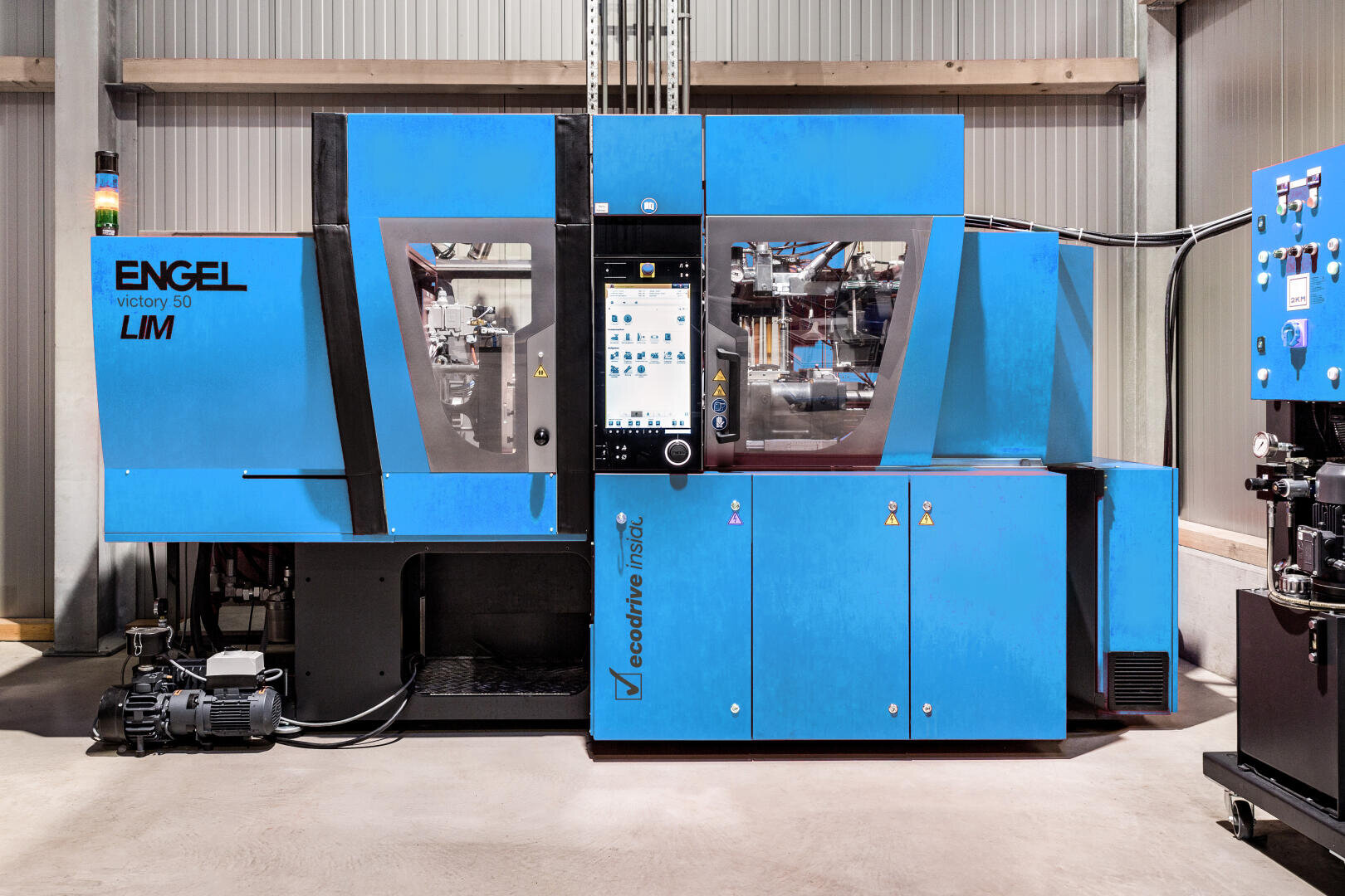 Eine große blaue ENGEL victory 50 LM Spritzgießmaschine steht in einem industriellen Umfeld mit einem zentralen Bedienfeld und sichtbaren Maschinenteilen hinter Glasscheiben.