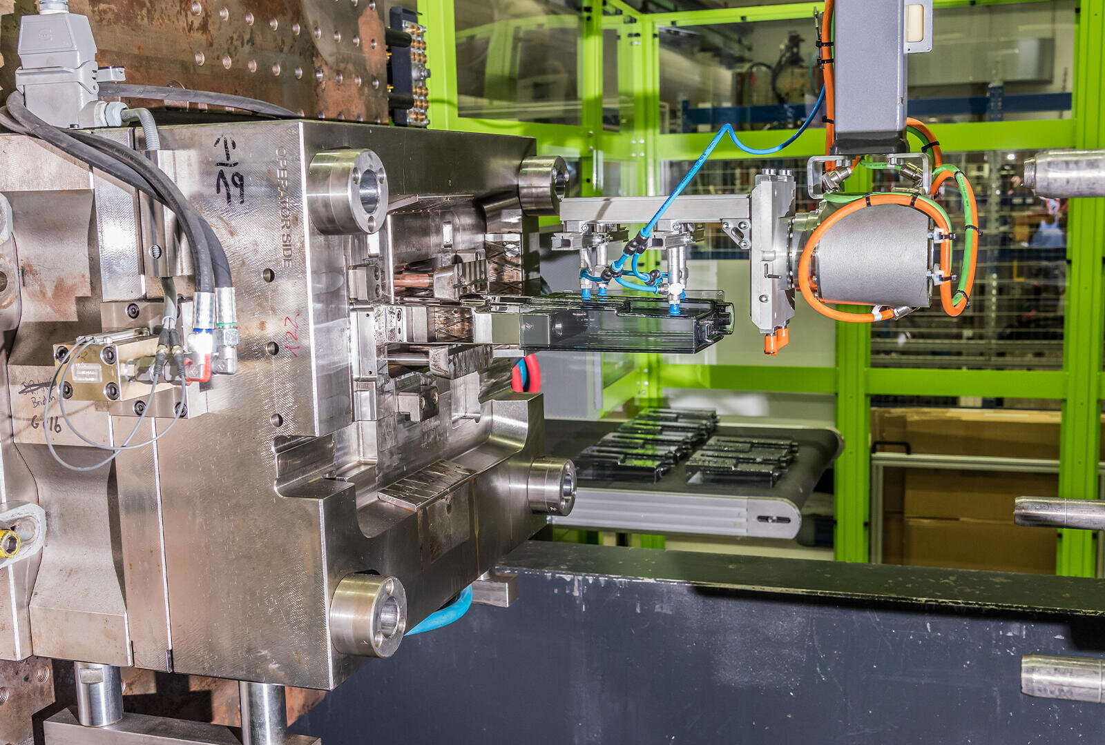 Ein Roboterarm bedient eine Spritzgießmaschine und bearbeitet ein Kunststoffteil in einer Produktionsstätte mit grün umrahmten Fenstern im Hintergrund.