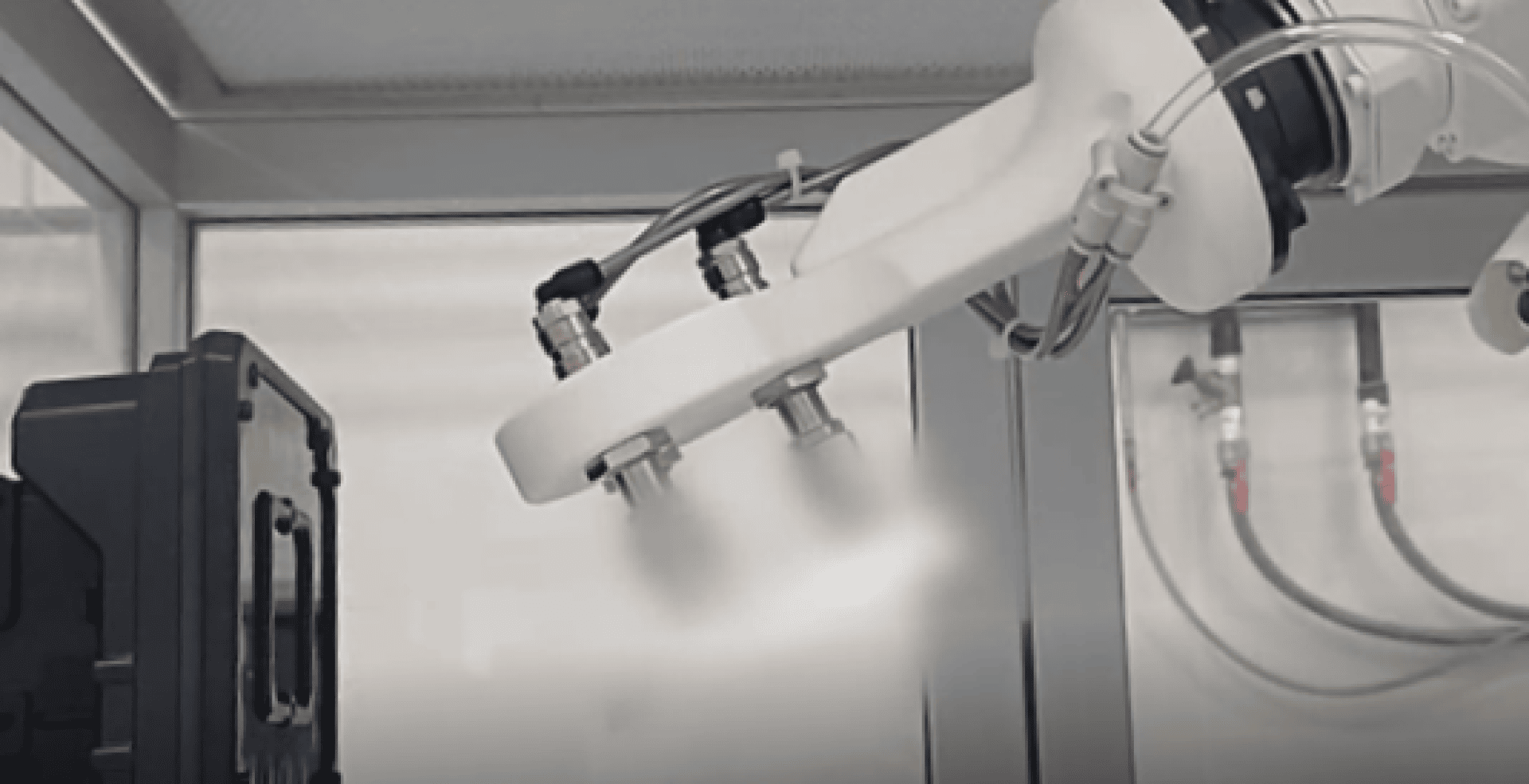 Un bras robotisé pulvérise un brouillard sur un composant à l'intérieur d'une enceinte industrielle, avec des câbles et des tuyaux visibles à l'arrière-plan.
