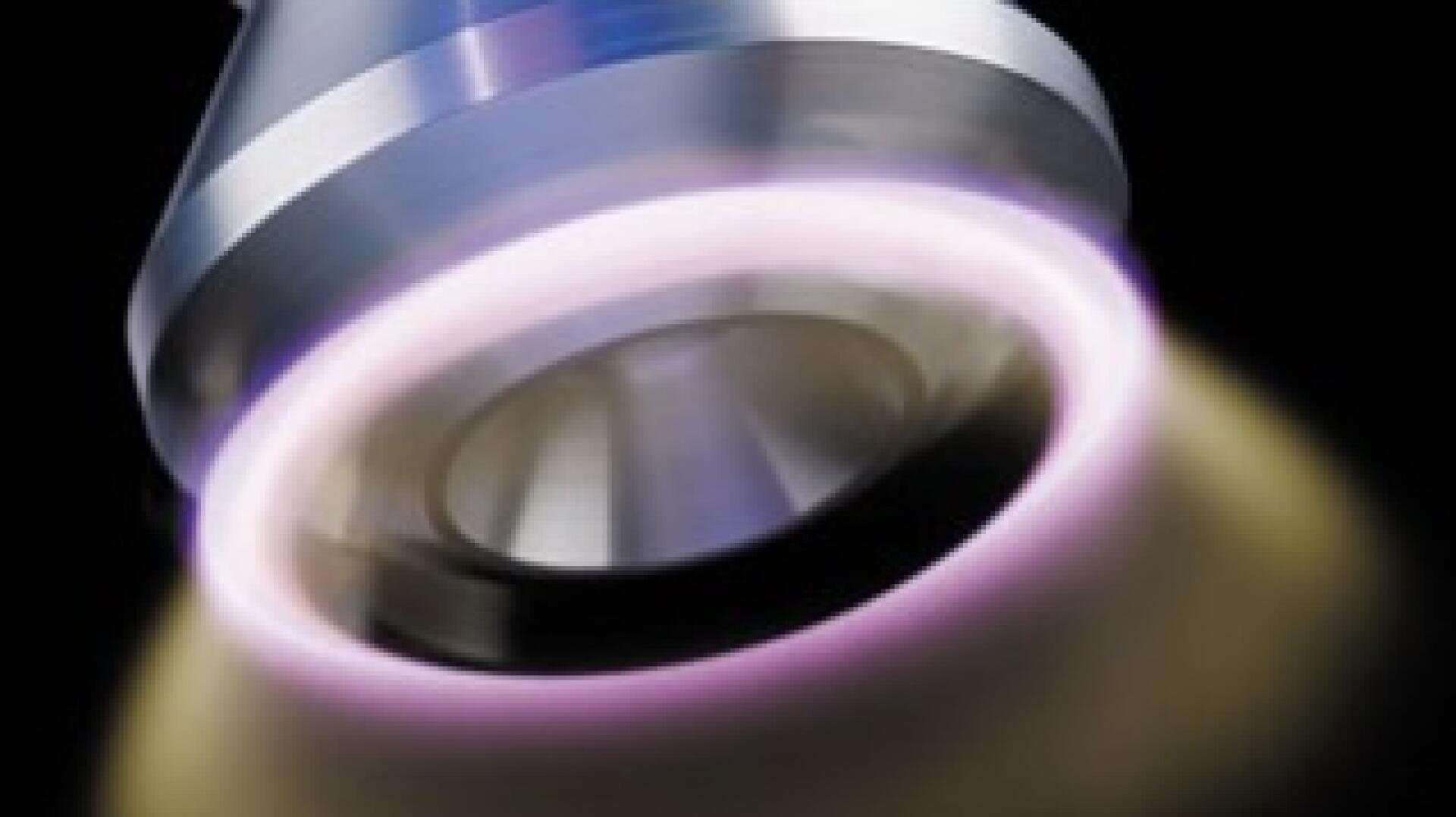 Gros plan sur une buse métallique émettant un courant concentré entouré d'une lumière violette circulaire incandescente, sur un fond sombre.