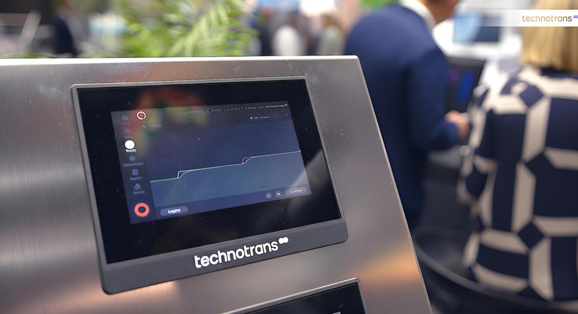 Nahaufnahme eines Touchscreen-Displays von technotrans, auf dem ein Liniendiagramm angezeigt wird, mit unscharfen Personen in Business-Kleidung im Hintergrund.