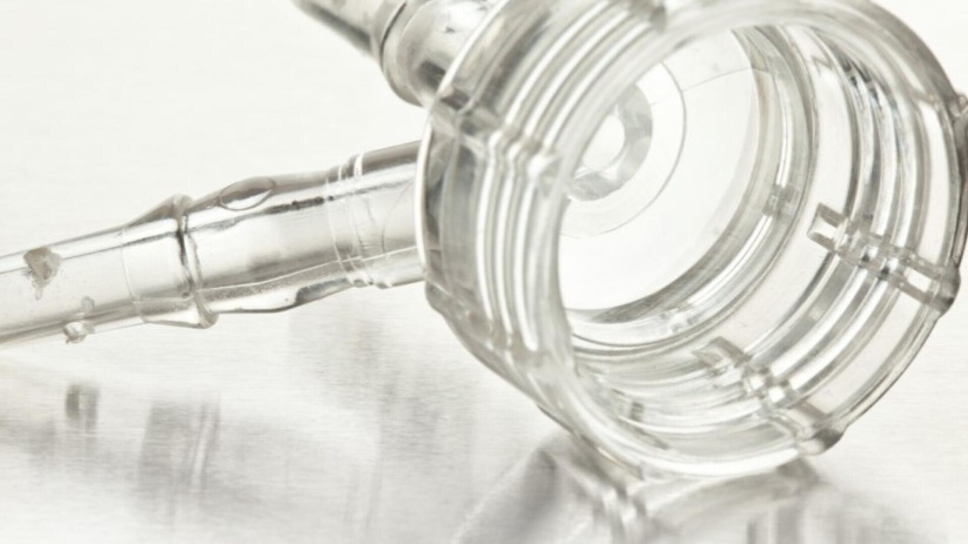 Gros plan sur un connecteur médical ou un adaptateur de tubulure en plastique transparent, posé sur une surface réfléchissante et présentant une ouverture circulaire cannelée et une extrémité effilée.