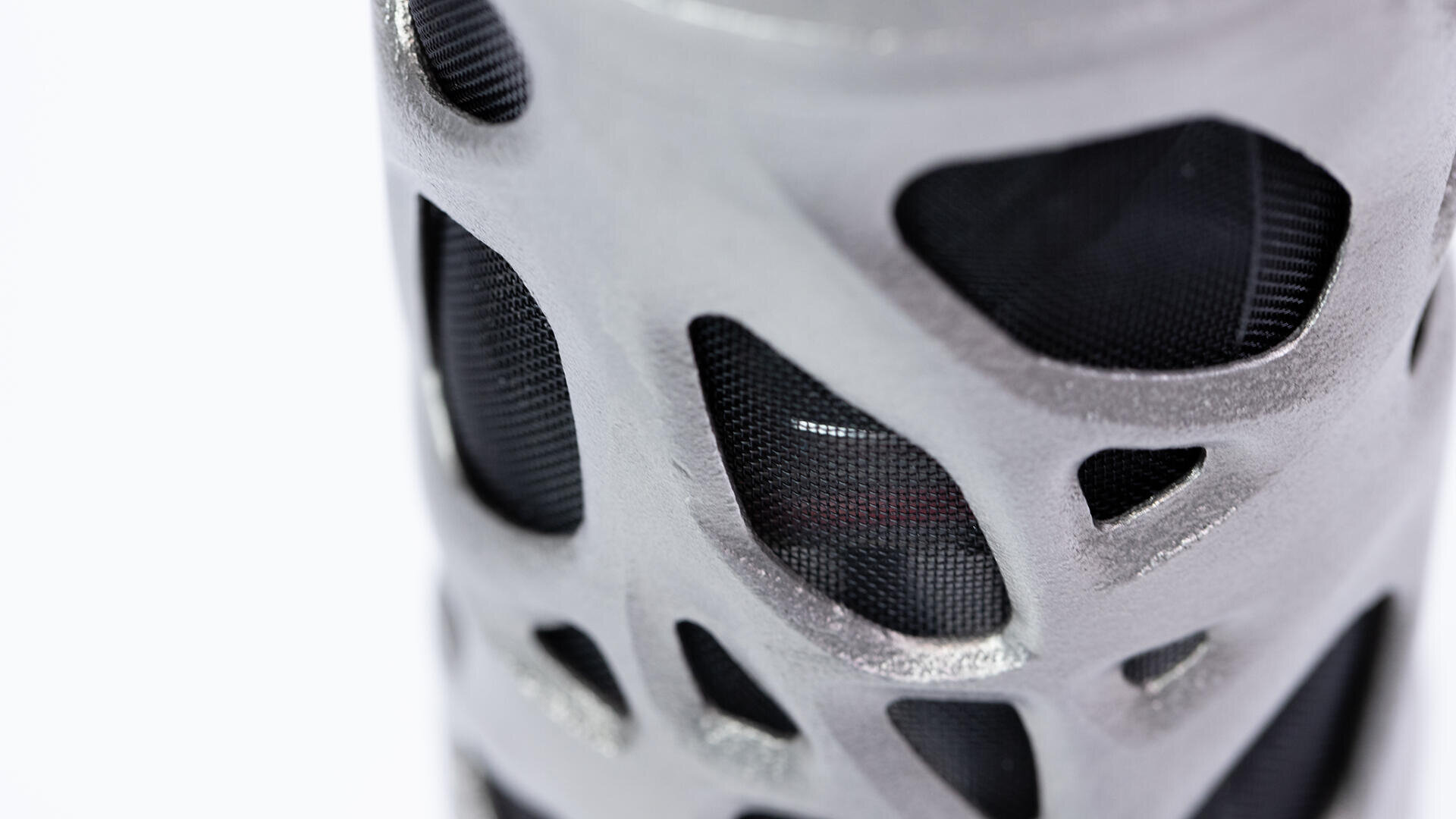 Nahaufnahme eines zylindrischen Objekts mit einer filigranen, silbernen, gitterartigen Außenhülle und einem dunklen, netzartigen Material, das durch die Öffnungen sichtbar ist. Der Hintergrund ist weiß und hebt die Details des Designs hervor.