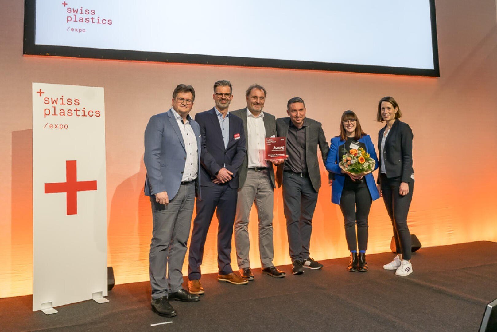 Sechs Personen stehen auf der Bühne der Swiss Plastics Expo und lächeln in die Kamera. Eine Person hält eine Trophäe, eine andere einen Blumenstrauß in der Hand. Auf der linken Seite ist ein Schild mit dem Logo der Veranstaltung zu sehen.