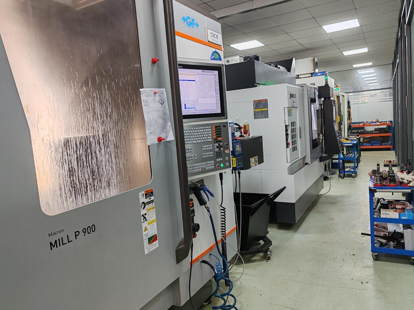 Eine Reihe von CNC-Fräsmaschinen in einer gut beleuchteten Industriehalle, mit sichtbaren Bedienfeldern und Werkzeugen. Die nächstgelegene Maschine mit der Bezeichnung MILL P 900 hat ein Fenster mit Kondenswasser, und neben den Maschinen stehen Wagen mit Teilen.