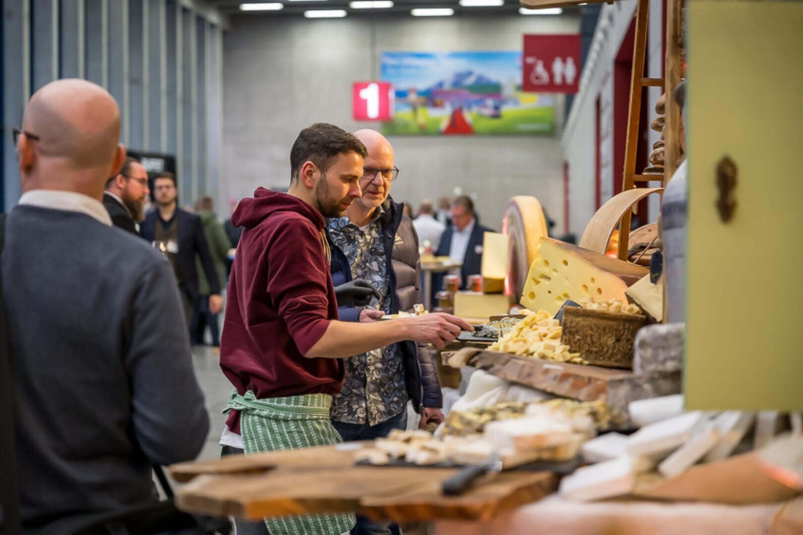 Plusieurs personnes se tiennent devant un comptoir à fromage dans un marché couvert, goûtent et choisissent des fromages. L'activité est intense, car les meules de fromage et les morceaux coupés sont exposés sur des surfaces en bois.